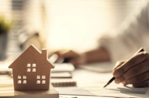 A imagem mostra uma casinha de madeira em primeiro plano, a mão de uma pessoa segurando uma caneta em cima de papéis, exemplificando bem cálculos para avaliar um investimento imobiliário. (Foto: Adobe Stock / Godshutter)