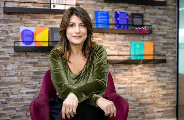 Mulher com cabelo castanho e blusa verde, sentada em poltrona roxa, olhando para frente. Parede de pedra com prateleiras ao fundo.