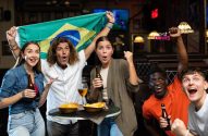 Torcedores brasileiros vibrando em um bar, com bandeira e bebidas.