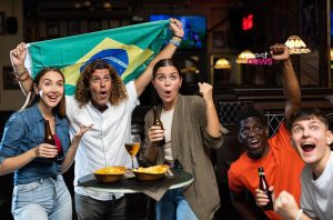 Torcedores brasileiros vibrando em um bar, com bandeira e bebidas.