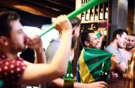 Torcedores brasileiros animados em um bar, um com a bandeira do Brasil e outro soprando uma buzina verde.