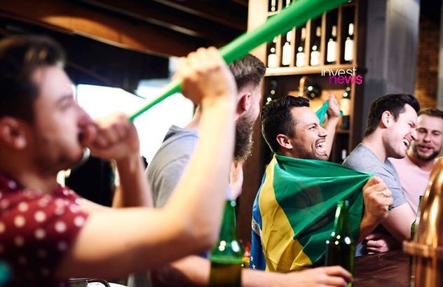 Torcedores brasileiros animados em um bar, um com a bandeira do Brasil e outro soprando uma buzina verde.