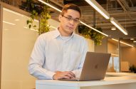 Homem de óculos olha para a câmera, com as mãos no teclado de um notebook, apoiado em um balcão de escritório.