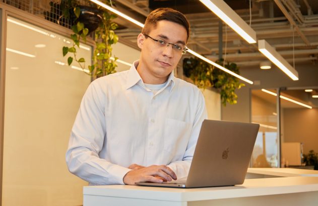 Homem de óculos olha para a câmera, com as mãos no teclado de um notebook, apoiado em um balcão de escritório.