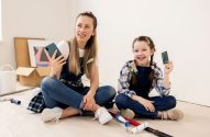 Mulher e garota em macacões, sentadas no chão. Mulher com celular, garota com cartão de crédito, sorrindo. Rolos de pintura próximos.