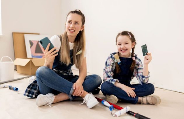 Mulher e garota em macacões, sentadas no chão. Mulher com celular, garota com cartão de crédito, sorrindo. Rolos de pintura próximos.