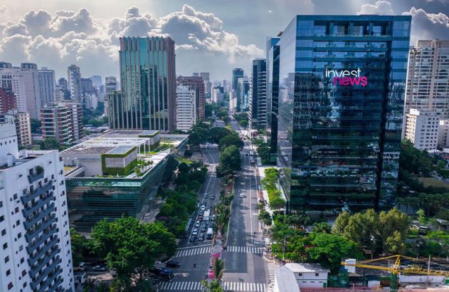 Vista aérea de avenida com tráfego, arranha-céus e vegetação. Um prédio exibe o logo 'invest news'.