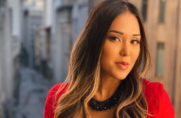 Retrato de mulher com cabelo escuro e mechas, blusa vermelha e colar. Ela olha para a câmera com fundo de cidade.