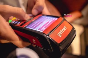 Mãos operam terminal de pagamento Getnet vermelho e preto, com tela e botões acesos.