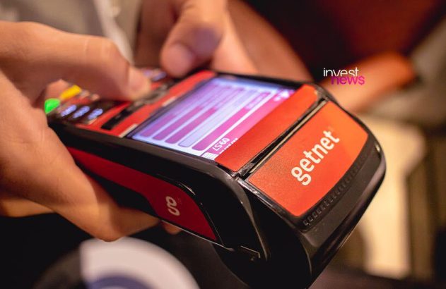 Mãos operam terminal de pagamento Getnet vermelho e preto, com tela e botões acesos.