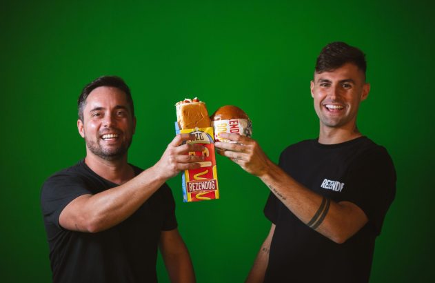 Dois homens sorrindo brindam com um cachorro-quente Rezendog e um hambúrguer Rezende em fundo verde.
