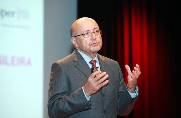 Homem careca de terno e óculos gesticula enquanto fala em palco com tela e cortinas vermelhas.