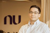 Homem de óculos sorrindo em camisa branca. Ao fundo, o logo roxo 'nu' (Nubank) aparece desfocado.