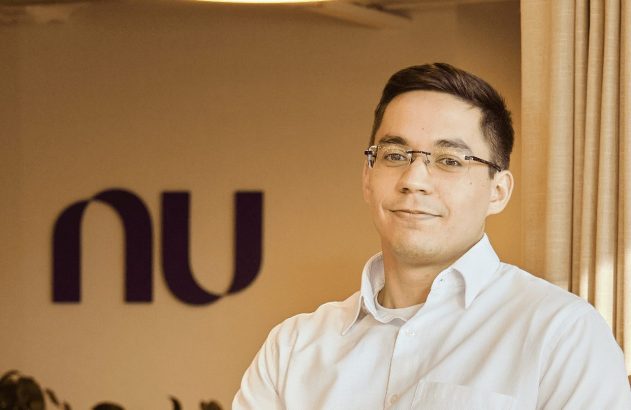 Homem de óculos sorrindo em camisa branca. Ao fundo, o logo roxo 'nu' (Nubank) aparece desfocado.