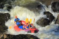 Pessoas em um bote de rafting vermelho e preto descem corredeiras turbulentas, entre água branca e rochas.