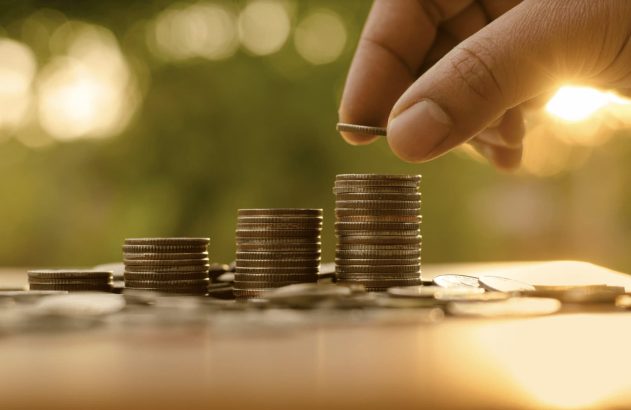 A imagem mostra a mão de uma pessoa branca adicionando uma moeda em cima de uma pilha de moedas. (Foto: Shutterstock / Singkham)