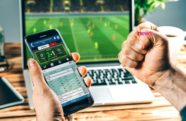 Pessoa usando celular com app de apostas de futebol, assistindo jogo em laptop; punho com 'news'.