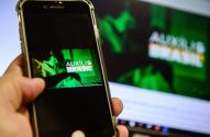 Mão segura smartphone exibindo 'Auxílio Brasil' em tela verde, com monitor similar ao fundo.