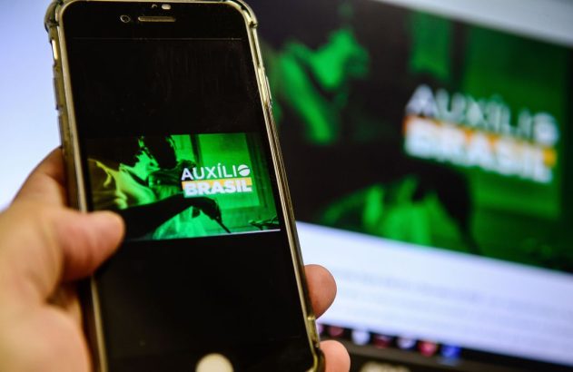 Mão segura smartphone exibindo 'Auxílio Brasil' em tela verde, com monitor similar ao fundo.
