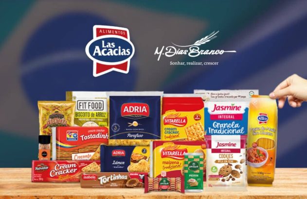 Mão segura macarrão Las Acacias entre produtos M.Dias Branco em mesa. Fundo com bandeira do Brasil.
