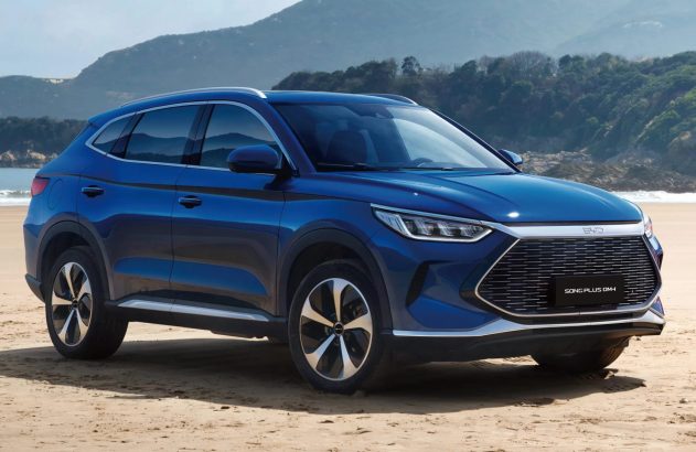 SUV azul BYD Song Plus DM-i em praia arenosa, com montanhas e oceano ao fundo. Vista frontal-lateral do veículo.