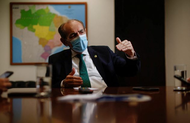 Homem de máscara azul, sentado à mesa, gesticula. Mapa do Brasil ao fundo.