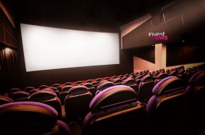 Interior de cinema vazio com tela grande e logo 'invest news'.