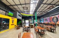 Amplo bar/restaurante com mesas, contêineres coloridos (amarelo 'Wine Bar'), murais e decoração brasileira.