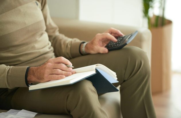 Pessoa sentada com calculadora e caderno, fazendo contas e anotações. Papéis na lateral.