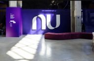 Interior de escritório roxo com logo 'nu', banco e sinalização direcional. Sombra no chão de concreto e pessoa perto de catraca.