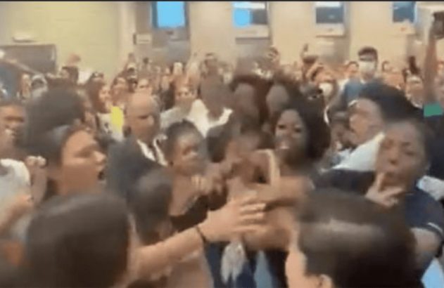 Mulheres em briga física no centro de uma multidão em ambiente interno. Pessoas observam e filmam.