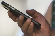 Mão de adulto com pele escura segura smartphone preto. Botões laterais do aparelho visíveis.