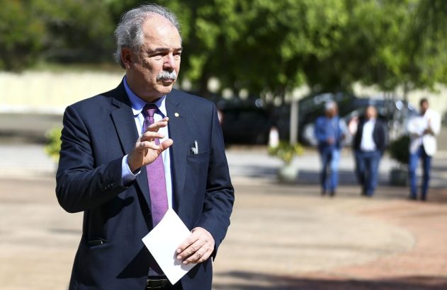 Homem de terno e bigode gesticula e segura papel. Fundo com pessoas e árvores desfocadas.