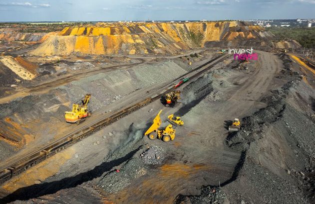 Operação de mineração a céu aberto: escavadeiras carregam trem e caminhões basculantes. Pilhas de minério ao redor.
