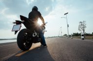 Motociclista com capacete aguarda em estrada sobre a água, com luz solar intensa.