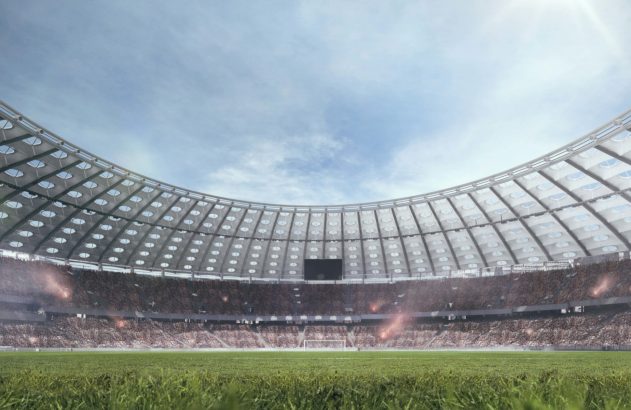 Vista de campo de futebol em estádio cheio de torcedores, com cobertura curva e céu claro. Sinalizadores acendendo.