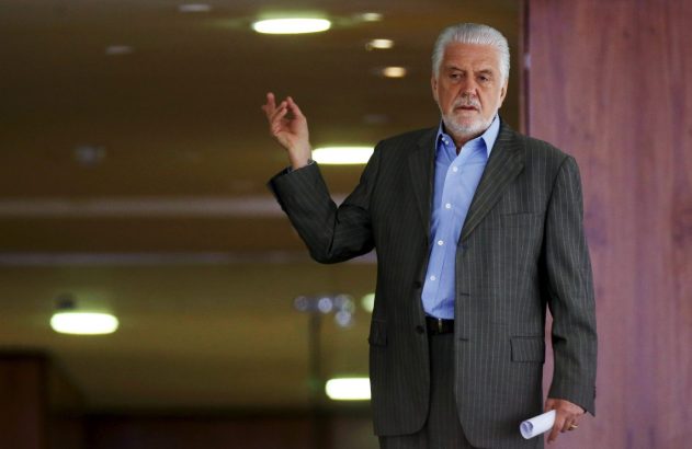 Homem de cabelo grisalho e barba, de terno e camisa azul, gesticula com a mão e segura um papel enrolado.