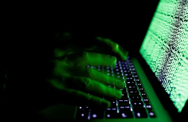 Mãos digitando em teclado de laptop com tela verde exibindo código binário, em ambiente escuro.