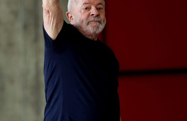 Homem com barba branca e camiseta azul levanta o braço direito em saudação, olhando para a frente. Fundo dividido.