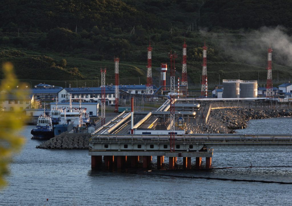 Terminal portuário industrial com longo píer, tubulações, tanques e torres. Barcos atracados e montanhas ao fundo.