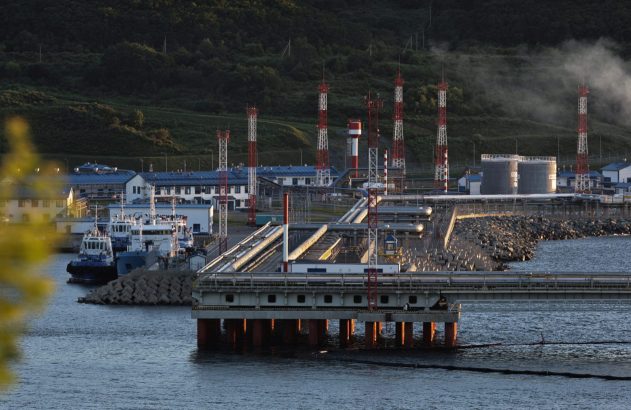 Terminal portuário industrial com longo píer, tubulações, tanques e torres. Barcos atracados e montanhas ao fundo.