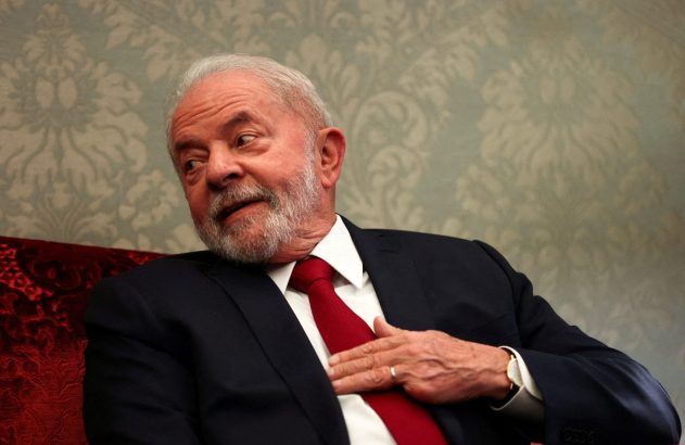 Homem idoso de barba branca, terno escuro e gravata vermelha, sentado, com a mão sobre o peito e olhando para a esquerda.