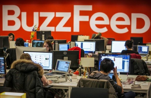 Escritório BuzzFeed. Pessoas trabalham em computadores. Logo 'BuzzFeed' grande em parede vermelha.