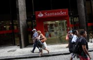 Pessoas caminham em calçada de rua urbana com o letreiro vermelho do banco Santander.