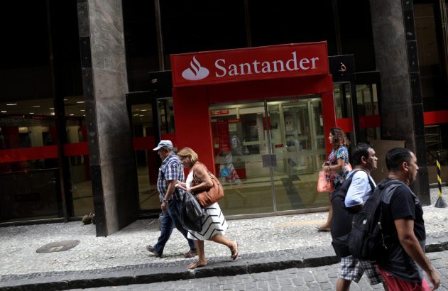 Pessoas caminham em calçada de rua urbana com o letreiro vermelho do banco Santander.