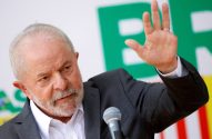 Lula, de barba e terno, fala ao microfone com a mão esquerda levantada e anel. Fundo desfocado verde, amarelo e vermelho.