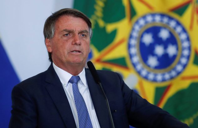Jair Bolsonaro, de terno, discursa ao microfone com a bandeira do Brasil desfocada ao fundo.