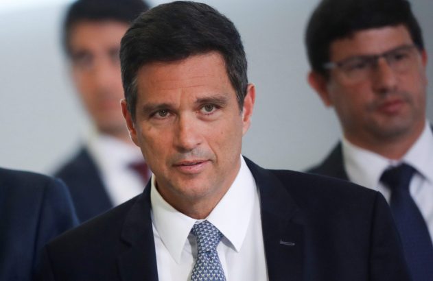 Close-up de homem de terno olhando para frente, com dois homens desfocados ao fundo.