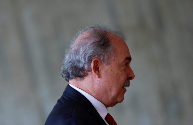 Homem de perfil com cabelo grisalho e bigode, usando terno escuro, camisa branca e gravata vermelha.