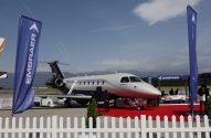 Jato Embraer Praetor 600 em exibição ao ar livre com carpete vermelho, bandeiras Embraer e montanhas ao fundo.